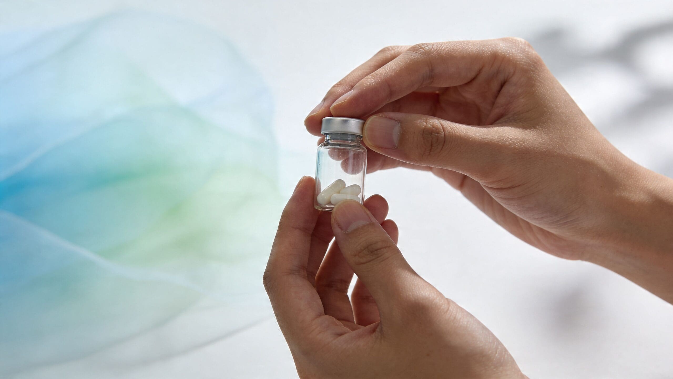 A pair of hands holding a small glass vial containing several white pills against a soft background.