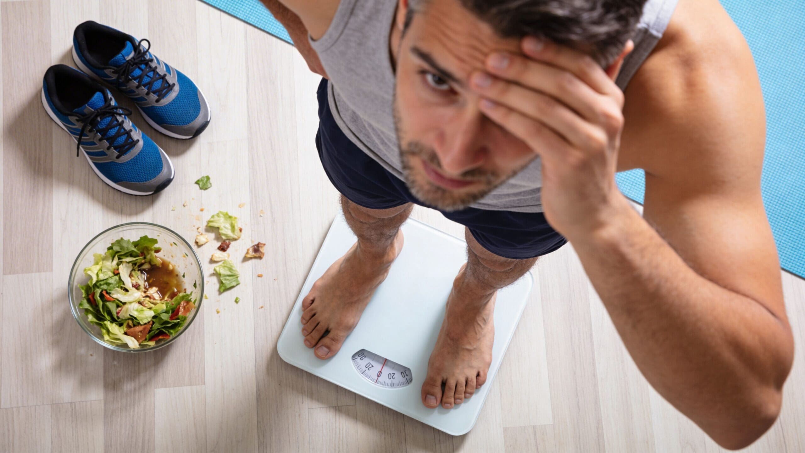 A man looking frustrated while standing on a weight scale next to a bowl of salad.