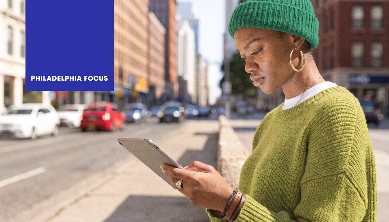 A woman wearing a green beanie uses a digital tablet on a sunny city street corner.
