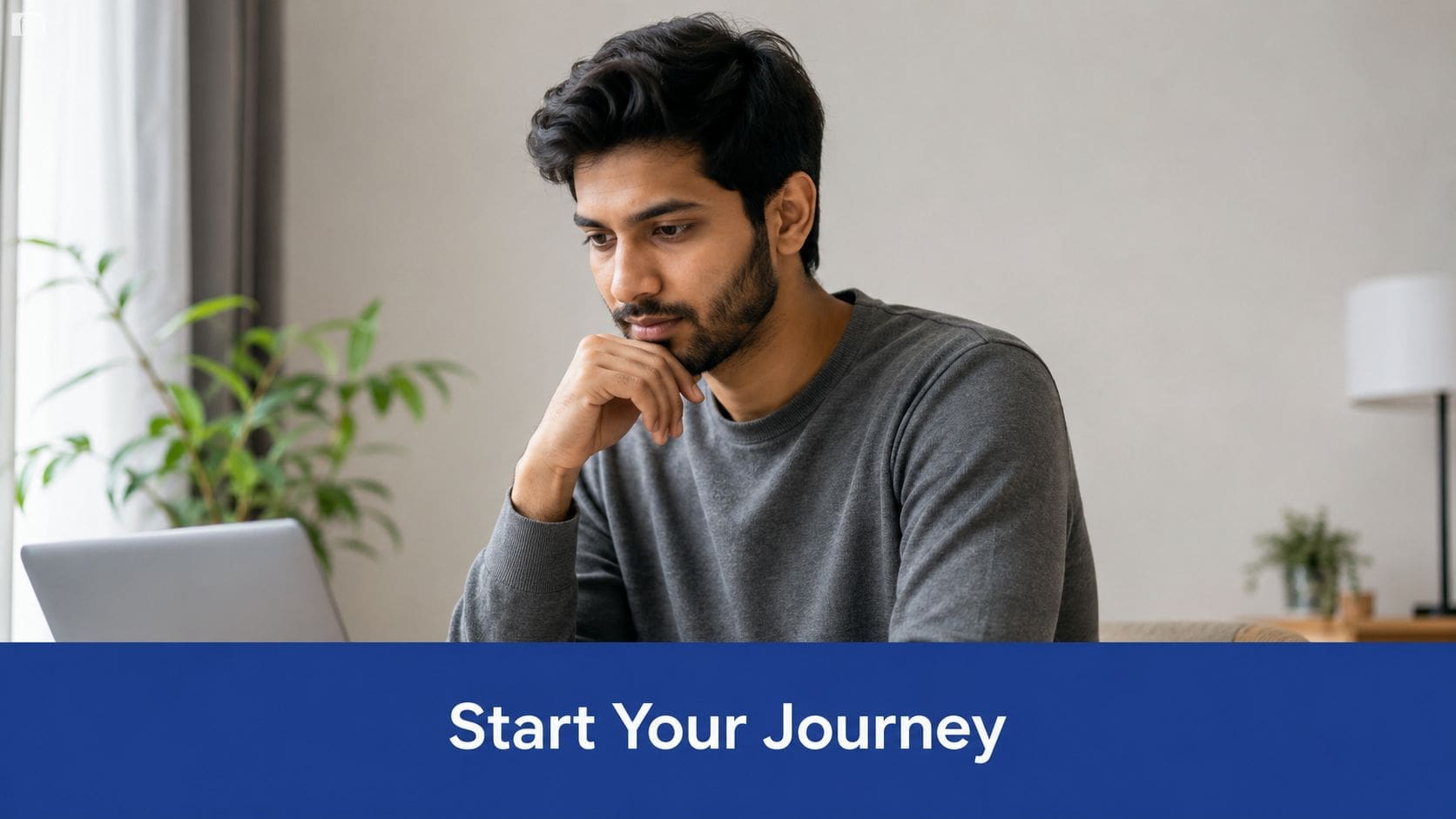 A man looking thoughtfully at his laptop screen while sitting at home with a plant nearby.
